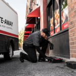 Pest-control technician kneeling by a sidewalk toolbox outside a restaurant, preparing equipment beside a white BITE-AWAY van.